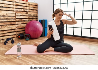 Middle age woman sitting on training mat at the gym using smartphone very happy and smiling looking far away with hand over head. searching concept.  - Powered by Shutterstock