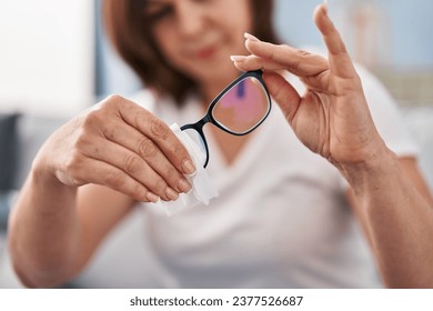 Middle age woman sitting on sofa cleaning glasses at home - Powered by Shutterstock