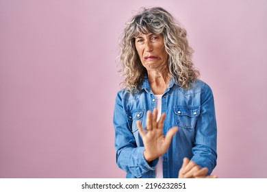 Middle age woman standing over pink background disgusted expression, displeased and fearful doing disgust face because aversion reaction. with hands raised 