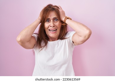 Middle age woman standing over pink background crazy and scared with hands on head, afraid and surprised of shock with open mouth  - Powered by Shutterstock