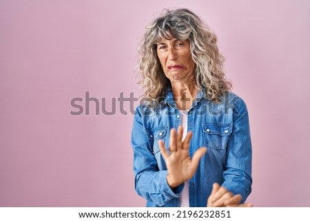 Middle age woman standing over pink background disgusted expression, displeased and fearful doing disgust face because aversion reaction. with hands raised 