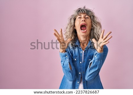 Middle age woman standing over pink background crazy and mad shouting and yelling with aggressive expression and arms raised. frustration concept. 