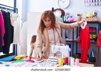 Middle age woman tailor holding t shirt writing on notebook at clothing factory - Shutterstock ID 2256893807