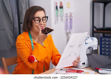 Middle age woman tailor talking on smartphone looking clothing design at tailor shop - Shutterstock ID 2256924127