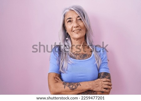 Middle age woman with tattoos standing over pink background happy face smiling with crossed arms looking at the camera. positive person. 