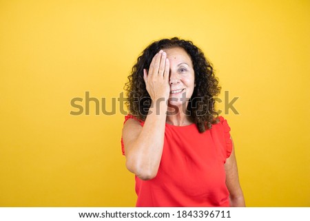 Middle age woman wearing casual shirt standing over isolated yellow background covering one eye with hand, confident smile on face and surprise emotion.