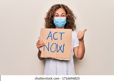 Middle Age Woman Wearing Coronavirus Protection Mask Holding Act Now Protest Cardboard Pointing Thumb Up To The Side Smiling Happy With Open Mouth