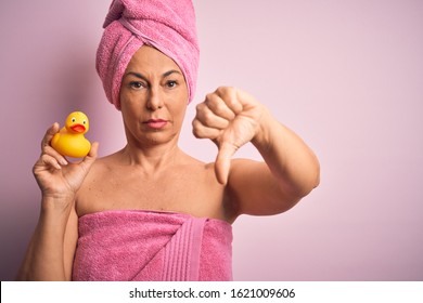 Middle age woman wearing pink bath towel from beauty body care holding rubber yellow duck with angry face, negative sign showing dislike with thumbs down, rejection concept