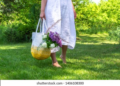 Middle Age Woman In White Simple Linen Dress Stays Barefoot On The Grass In Beautiful Garden And Holds Knitted White-yellow Bag With Flowers Bouquet  Inside, Placeholder 