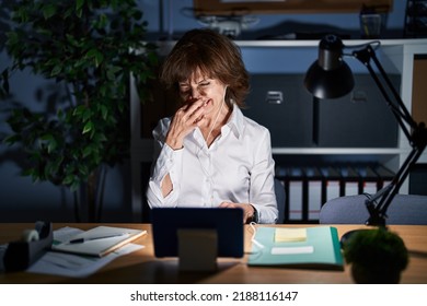 Middle Age Woman Working At The Office At Night Smelling Something Stinky And Disgusting, Intolerable Smell, Holding Breath With Fingers On Nose. Bad Smell 