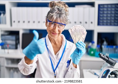 Middle Age Woman Working At Scientist Laboratory Holding Dollars Smiling Happy And Positive, Thumb Up Doing Excellent And Approval Sign 