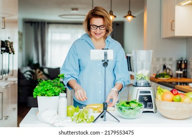 Middle Age Women Is Blogging For Her Wellness Lifestyle Channel About Healthy Balanced Living In The Kitchen Of Her Home. Creating Video Content For Social Media With Phone Camera On A Tripod