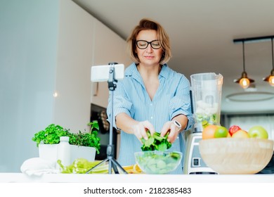 Middle Age Women Is Blogging For Her Wellness Lifestyle Channel About Healthy Balanced Living In The Kitchen Of Her Home. Creating Video Content For Social Media With Phone Camera On A Tripod