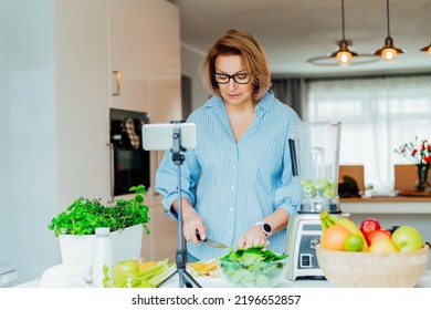 Middle Age Women Is Blogging For Her Wellness Lifestyle Channel About Healthy Balanced Living In The Kitchen Of Her Home. Creating Video Content For Social Media With Phone Camera On A Tripod