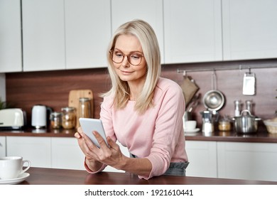Middle aged 50 years old woman using apps ordering buying food on smartphone sitting in kitchen at home. Mature older lady holding mobile phone texting messages, browsing online services. - Shutterstock ID 1921610441