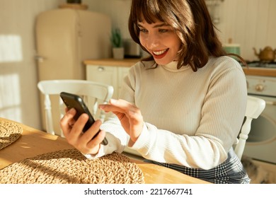 Middle Aged 50 Years Old Woman Using Apps Ordering Buying Food On Smartphone Sitting In Kitchen At Home. Mature Older Lady Holding Mobile Phone Texting Messages, Browsing Online Services.