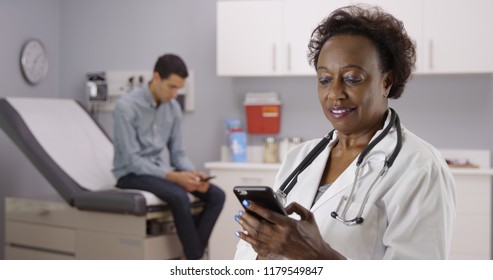 Middle Aged African Doctor Using Mobile Phone To Text While Male Patient Waits