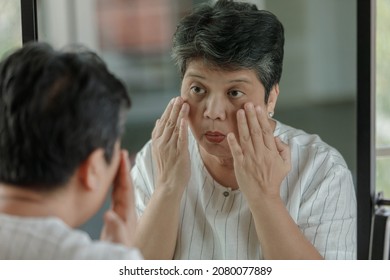 Middle aged Asian female touching face and checking under eye puffiness and wrinkle while looking at mirror at home