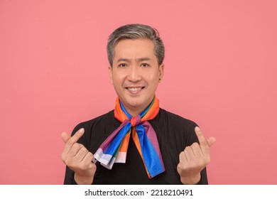 Middle Aged Asian Gay Man With Gray Hair  Wearing A Black Turtleneck Is Smiling Make A Mini Heart Symbol And Tie A Rainbow Flag Scarf Isolated On Pink Bakcground. Concept Mature Gay Man, Lgbtq.