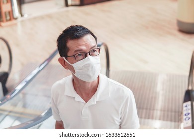 Middle Aged Asian Man Wearing Glasses And Medical Face Mask On Escalator, Post Covid-19 Coronavirus  Pandemic, New Normal, Air Pollution And Health, Reopening  Concept