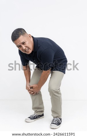 A middle aged asian man winces in pain. Bending down clutching his bum knee. Isolated on a white background.