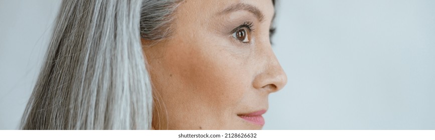 Middle Aged Asian Woman With Beautiful Straight Hoary Hair Poses On Light Grey Background In Studio Closeup Side View. Mature Beauty Lifestyle