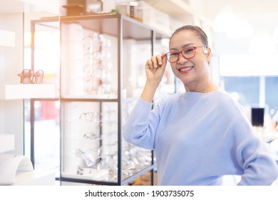 Middle Aged Asian Woman Choosing Spectacles Glasses In Optician Store. Used Correct Or Assist Defective Eyesight, Smiling And Looking At Camera.