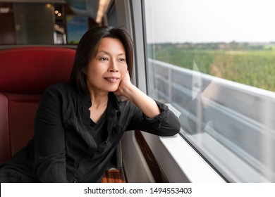 Middle Aged Asian Woman Looking Out The Window At Landscape View Sitting On Train Commute Travel Lifestyle. Mature Chinese Passenger Traveling Smiling Relaxing Thinking Of Holiday In Contemplation.