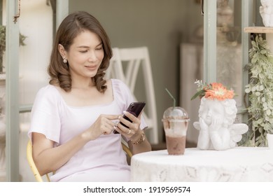 Middle Aged Asian Woman Sitting In Coffee Shop And Using Smartphone With Happiness. Business Owner And Freelance Lifestyle Concept.