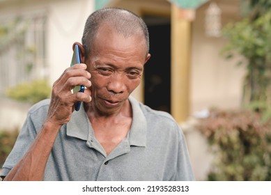 A Middle Aged, Balding Filipino Male In His Early 60s Conversing On His Cellphone. Candid Scene Outside His Home.