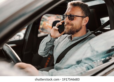 Middle Aged Bearded Man With Eyeglasses Driving His Car And Having Phone Conversation. It's Unsafe And Dangerous To Do It In Traffic.