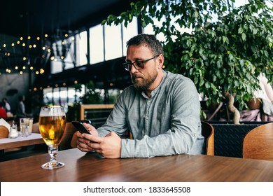 Middle aged bearded man sitting in a bar and using smart phone for hanging on social media sites. In front of him on a table is a cold fresh beer. - Powered by Shutterstock