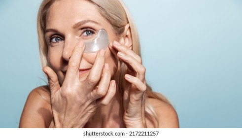 Middle aged beauty woman, applies under eye patches from aging wrinkles, puffiness and dark circles, standing over blue background - Powered by Shutterstock