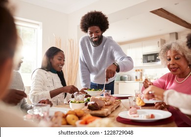 Middle Aged Black Man Carving And Serving Meat At Sunday Family Dinner With His Partner, Kids And Their Grandparents, Front View