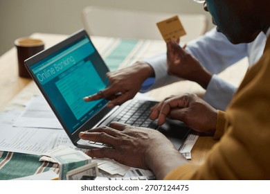 Middle aged Black man using laptop for online tax payment while middle aged Black woman holding credit card and pointing at computer screen, tax documents and cash on table