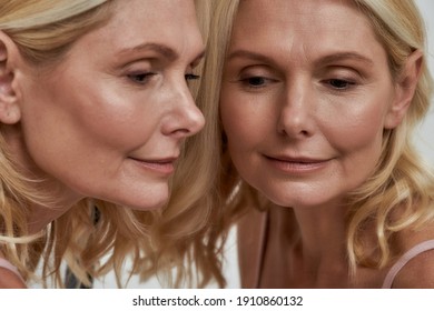 Middle Aged Blond Caucasian Woman Reflecting In Mirror With Eyes Looking Down And Smile Against Light Background. Authentic Woman Beauty Concept