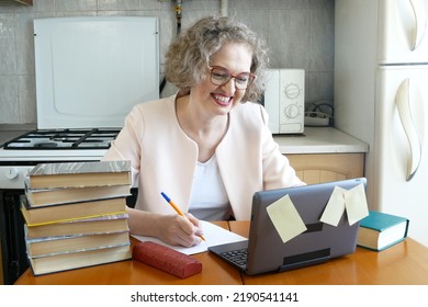 Middle Aged Blond Woman Watching Professional Training Class Or Online Webinar On Laptop In Kitchen With Paper Book Pile In Desk, Woman With Computer Remote Working, Distance Learning From Home Office