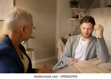 Middle Aged Blonde Female Mentor Or Coach Talking To Young Male, Teaching Intern Having Business Conversation With Workmate. Mature Woman Helping Man Colleague, Discussing New Project In Office