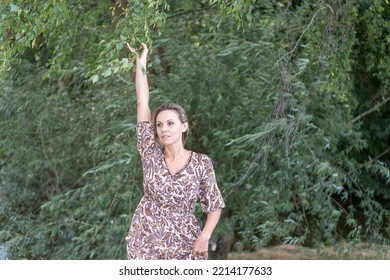 Middle Aged Blonde Woman Stretching One Arm Up With A Sensual Look At The Camera Outside In Front Of Green Trees