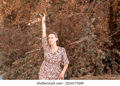 Middle Aged Blonde Woman Stretching One Arm Up With A Sensual Look At The Camera Outside In Front Of Autumn Trees