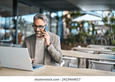 Middle aged business man talking on mobile phone hybrid working on laptop sitting outside office. Mature businessman making call on cellphone speaking to client using computer outdoors. Copy space. - Powered by Shutterstock