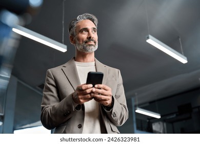 Middle aged business man wearing suit using cellphone looking away in office. Mature older businessman executive leader wearing suit holding mobile phone at work standing with smartphone in hands. - Powered by Shutterstock