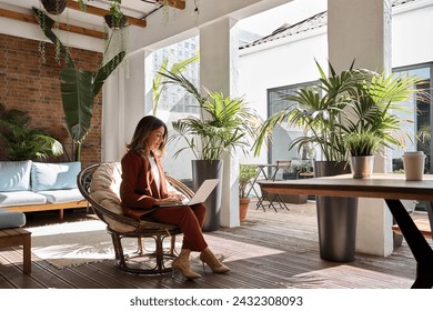 Middle aged business woman using laptop sitting in chair in sunny green office. Busy professional mature female entrepreneur or executive wearing suit holding computer on lap working. Candid photo. - Powered by Shutterstock