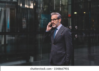 Middle Aged Businessman With Beard In Stylish Dark Business Suit Talking On Phone Near Office Building Putting Hand In Pocket In New York