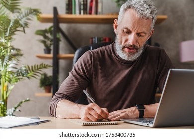 Middle aged businessman in casual clothes making notes in notepad while sitting in his office