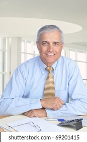 Middle Aged Businessman Seated At His Desk In Modern Office Setting. Man Is Wearing A Blue Dress Shirt And Yellow Tie With No Jacket. Vertical Format