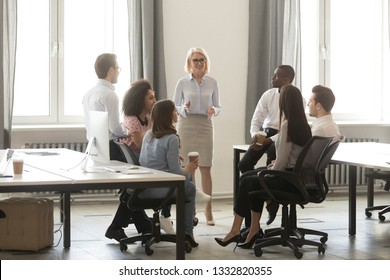 Middle aged businesswoman boss mentor training interns employees in modern office space, female business leader coach speaker teach instructing multicultural workers sales team at corporate meeting
