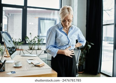 Middle aged businesswoman holding her stomach in discomfort while standing in front of a desk in an office setting. - Powered by Shutterstock