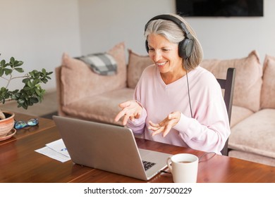 Middle aged businesswoman using earphone while sitting behind her laptop and having discussion and online meeting in video call. Gray haired businesswoman working from home. Home office