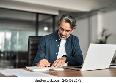 Middle aged busy professional businessman ceo writing notes learning online working in office. Mature male entrepreneur or executive sitting at work desk using laptop computer technology.